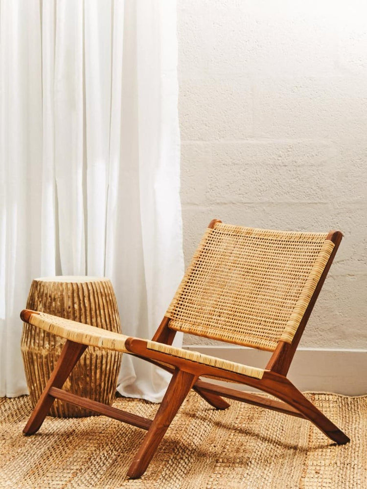 A wooden lounge chair with a natural rattan seat and backrest, positioned at an angle on a light wooden floor. A woven basket is placed nearby, and sheer white curtains hang in the background, allowing soft light to filter through