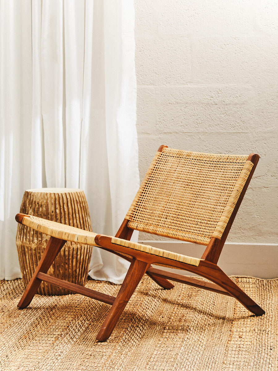 A wooden lounge chair with a natural rattan seat, featuring a minimalist design and angled backrest, positioned on a light-colored floor. A woven basket is placed beside the chair, and soft, sheer curtains hang in the background, allowing natural light to filter through
