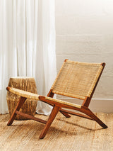 A wooden lounge chair with a natural rattan seat, featuring a minimalist design and angled backrest, positioned on a light-colored floor. A woven basket is placed beside the chair, and soft, sheer curtains hang in the background, allowing natural light to filter through