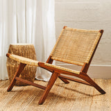 A wooden lounge chair with a natural rattan seat and backrest, featuring a minimalist design and angled legs. Next to the chair is a round, straw-like side table. The background includes light-colored curtains and a textured floor, creating a warm and inviting atmosphere