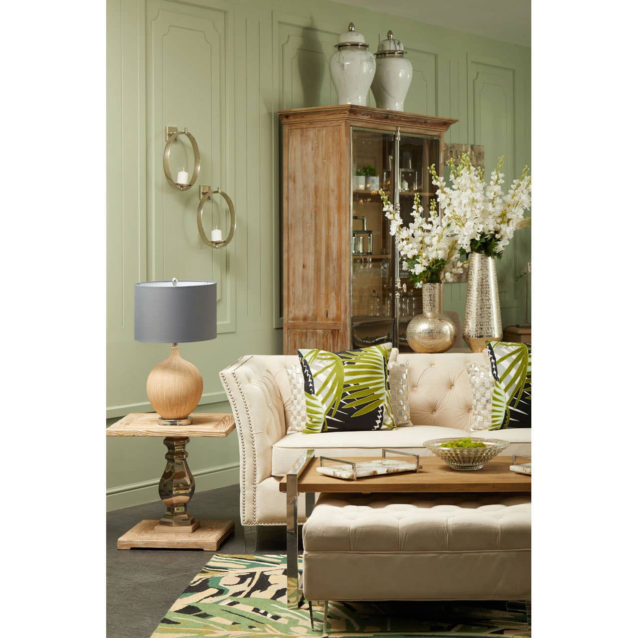 A white washed pine wood two-door cabinet stands against a soft green wall, showcasing its natural texture. The cabinet features glass doors, allowing visibility of the items inside, and is complemented by a cozy living room setting with a light-colored sofa adorned with green and patterned cushions. A wooden side table with a lamp sits beside the sofa, enhancing the warm ambiance of the space. Decorative vases and flowers are