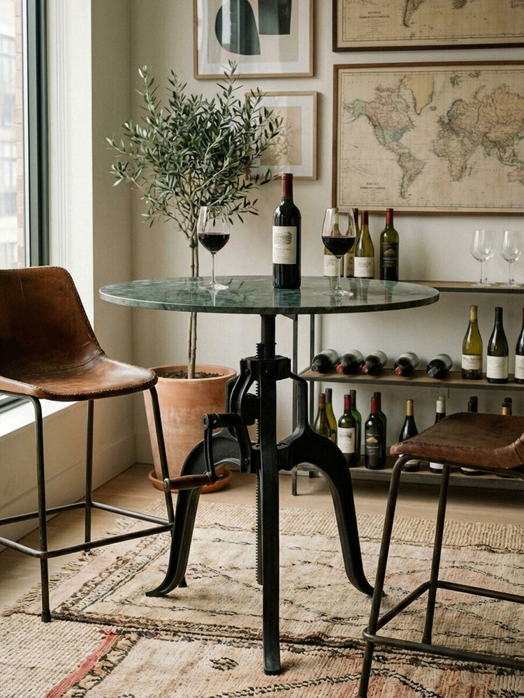 Small bar table featuring a round green marble top supported by a black metal base with a unique design. Two brown leather chairs are positioned beside the table, and a bottle of wine with two glasses sits atop the table. In the background, a collection of wine bottles is displayed on a shelf, and framed maps adorn the wall. A soft area rug is visible beneath the table and chairs, adding warmth to