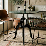 Small bar table featuring a round green marble top supported by a black metal base with a unique design. Two brown leather bar stools are positioned beside the table, and a bottle of wine with two glasses sits on the table. In the background, a shelf displays various wine bottles, and a decorative plant adds a touch of greenery to the scene. The setting is cozy and stylish, with a patterned rug under
