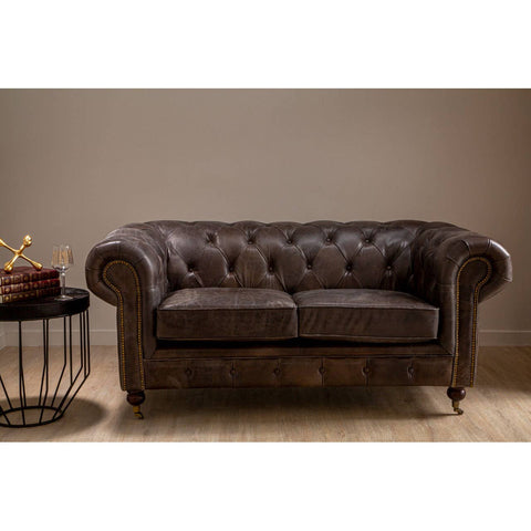 Dark grey brown leather Chesterfield 2 seat sofa with tufted backrest and rolled arms, positioned next to a round side table with a decorative bowl, set against a neutral wall and wooden floor