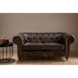 Dark grey brown leather Chesterfield 2 seat sofa with tufted backrest and rolled arms, positioned next to a round side table with a decorative bowl, set against a neutral wall and wooden floor