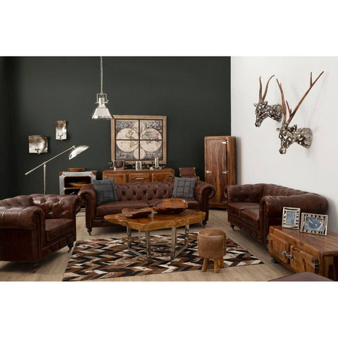 Dark grey brown leather Chesterfield 2 seat sofa positioned in a stylish living room setting, featuring a dark green accent wall, wooden furniture pieces, decorative antlers on the wall, and a cozy area rug. The sofa has a classic tufted design with rolled arms and is complemented by throw pillows