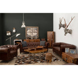 Dark grey brown leather Chesterfield 2 seat sofa positioned in a stylish living room setting, featuring a dark green accent wall, wooden furniture pieces, decorative antlers on the wall, and a cozy area rug. The sofa has a classic tufted design with rolled arms and is complemented by throw pillows