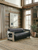 Black leather three-seat sofa with a modern design, featuring sleek armrests and a minimalist frame. The sofa is positioned in a cozy room with wooden walls, large windows allowing natural light, and a decorative plant in a pot nearby. A textured area rug covers the floor, enhancing the warm atmosphere of the space