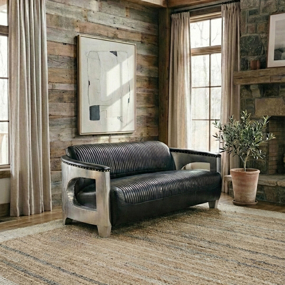 Black leather three-seat sofa with a modern design, featuring sleek metal arms and a minimalist aesthetic, placed in a cozy room with wooden walls, large windows, and a decorative plant nearby. A large mirror hangs on the wall, reflecting natural light into the space