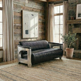 Black leather three-seat sofa with a modern design, featuring sleek metal arms and a minimalist aesthetic, placed in a cozy room with wooden walls, large windows, and a decorative plant nearby. A large mirror hangs on the wall, reflecting natural light into the space