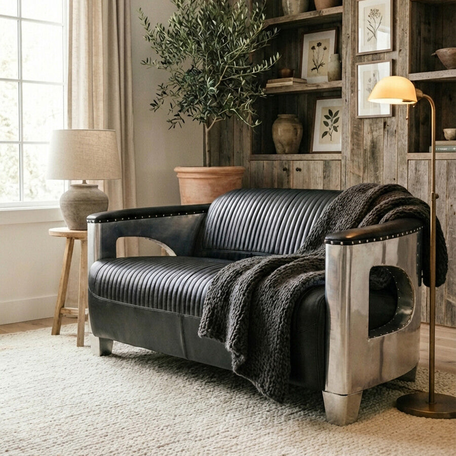 Black leather two-seat sofa positioned in a cozy living room setting, featuring a textured throw blanket draped over one arm. The background includes a wooden bookshelf with framed pictures, a potted plant, and a lamp on a side table, all complemented by natural light coming through a window. A soft area rug covers the floor