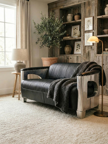 Black leather two-seat sofa positioned in a cozy living room setting, featuring a textured throw blanket draped over one arm. The background includes a wooden bookshelf with framed pictures, a potted plant, and a lamp on a side table, all complemented by natural light coming through a window. A soft area rug covers the floor