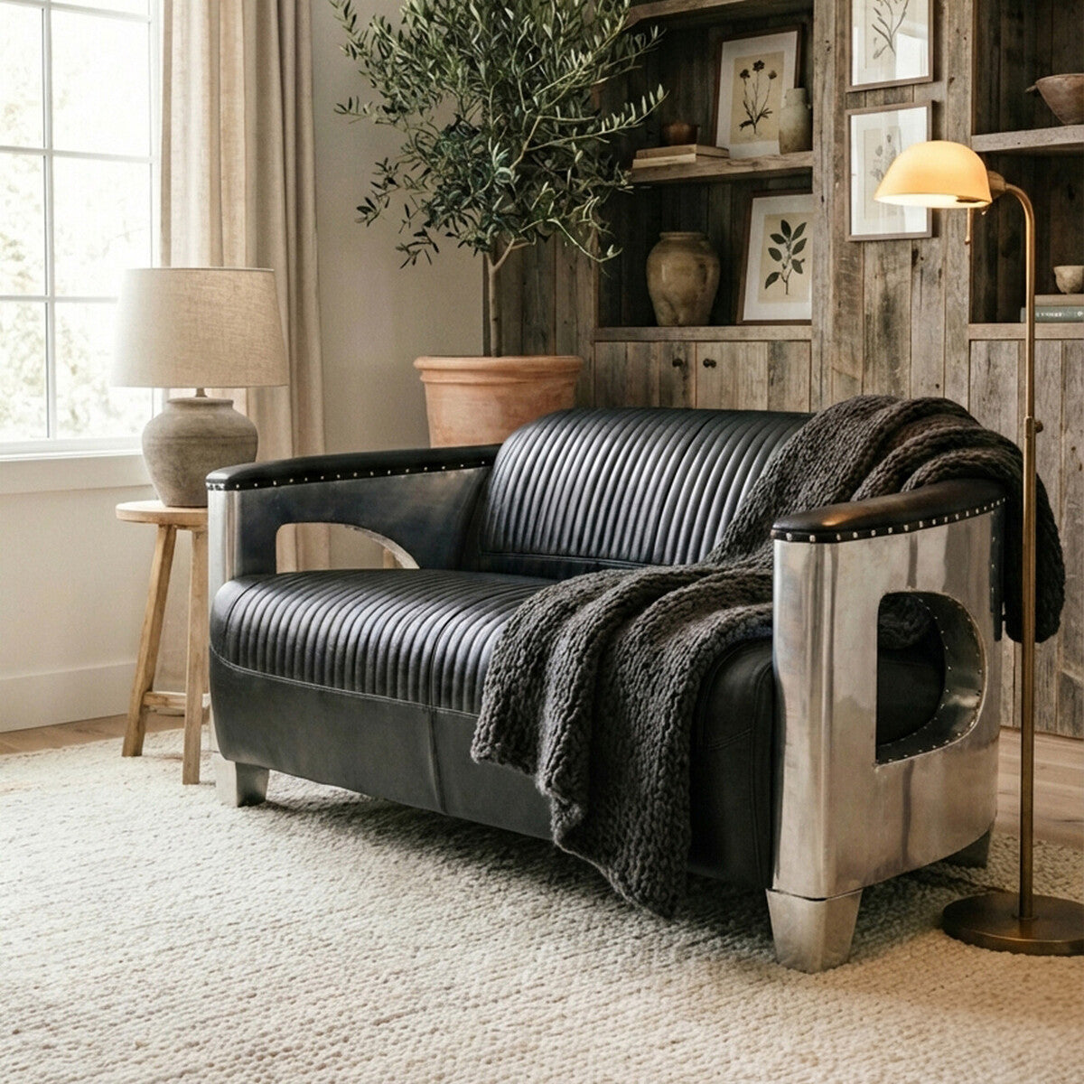 Black leather two-seat sofa with a modern design, featuring a sleek frame and plush cushions. A cozy black throw blanket is draped over one armrest. The sofa is positioned in a well-lit room with a large window, a stylish lamp, and a bookshelf in the background, creating a warm and inviting atmosphere