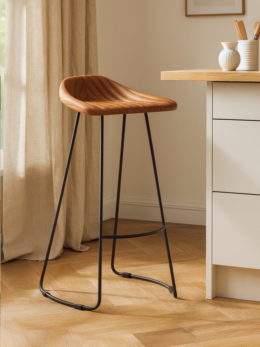 Light brown leather bar stool with a curved seat and a footrest, supported by sleek black metal legs, positioned next to a kitchen counter in a bright room with natural light and wooden flooring