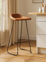 Light brown leather bar stool with a curved seat and a footrest, supported by sleek black metal legs, positioned next to a kitchen counter in a bright room with natural light and wooden flooring