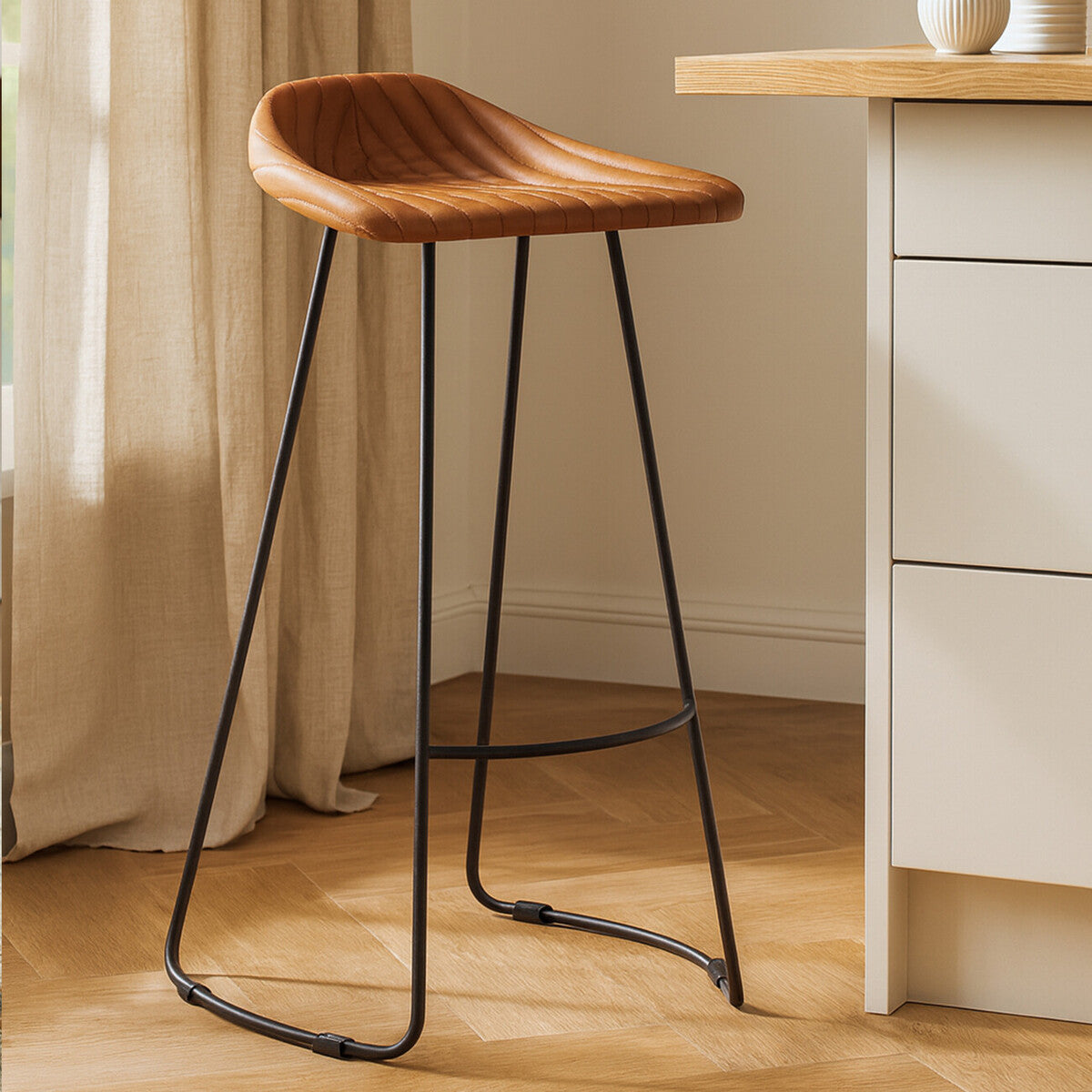 Light brown leather bar stool with a curved seat and a black metal frame, featuring a footrest, positioned next to a wooden countertop and soft curtains in a bright room