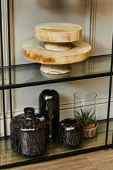A small wooden riser with a natural finish sits on a glass shelf, displaying two circular wooden pieces stacked on top of each other. Below, several dark glass vases and a small potted plant are arranged, creating an aesthetically pleasing display. The background features a light-colored wall, enhancing the warm tones of the wood and glass