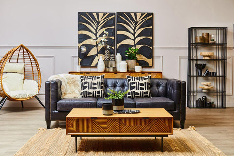 A small wooden riser placed on a coffee table in a modern living room setting. The riser features a natural wood finish and is adorned with decorative items, including a potted plant and a candle. In the background, a navy blue wall art piece with a leaf design complements the decor, while a cozy sofa with patterned cushions and a stylish shelving unit are also visible. A hanging chair adds a
