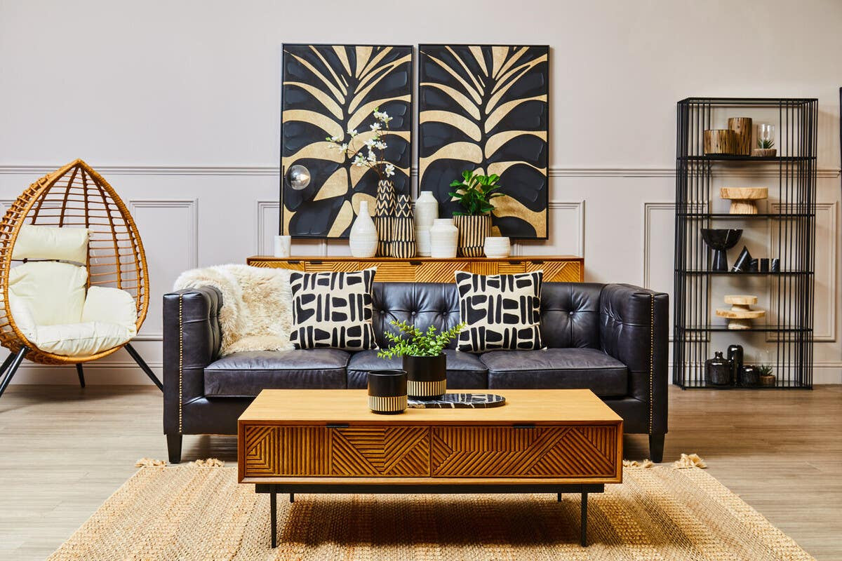 A small wooden riser placed on a coffee table in a modern living room setting. The riser features a natural wood finish and is adorned with decorative items, including a potted plant and a candle. In the background, a navy blue wall art piece with a leaf design complements the decor, while a cozy sofa with patterned cushions and a stylish shelving unit are also visible. A hanging chair adds a