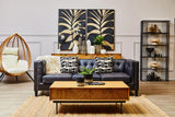 A small wooden riser placed on a coffee table in a modern living room setting. The riser features a natural wood finish and is adorned with decorative items, including a potted plant and a candle. In the background, a navy blue wall art piece with a leaf design complements the decor, while a cozy sofa with patterned cushions and a stylish shelving unit are also visible. A hanging chair adds a