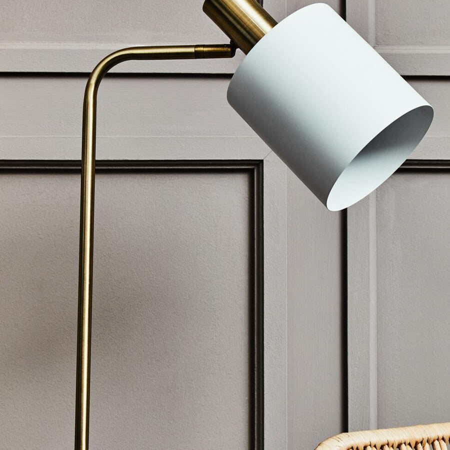 Brass floor lamp with white cylindrical shade beside rattan chair against panelled wall
