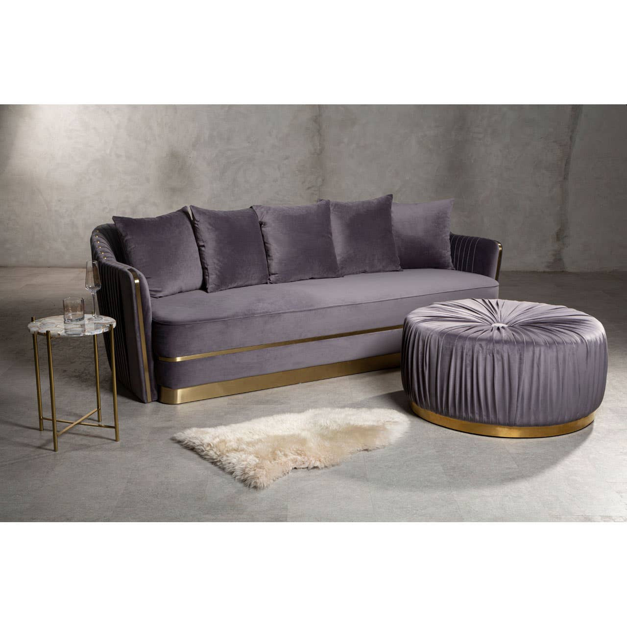 A round grey footstool with a tufted design sits next to a grey sofa with plush cushions. The footstool features a gold base, complementing the modern aesthetic of the living space. A small round side table and a light-colored rug are also visible in the scene