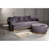 A round grey footstool with a tufted design sits next to a grey sofa with plush cushions. The footstool features a gold base, complementing the modern aesthetic of the living space. A small round side table and a light-colored rug are also visible in the scene