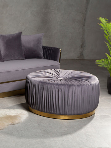 Round grey footstool with tufted upholstery and a gold circular base, placed next to a grey sofa with matching cushions, set against a textured wall and accompanied by a green plant in a pot
