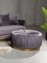 Round grey footstool with tufted upholstery and a gold circular base, placed next to a grey sofa with matching cushions, set against a textured wall and accompanied by a green plant in a pot