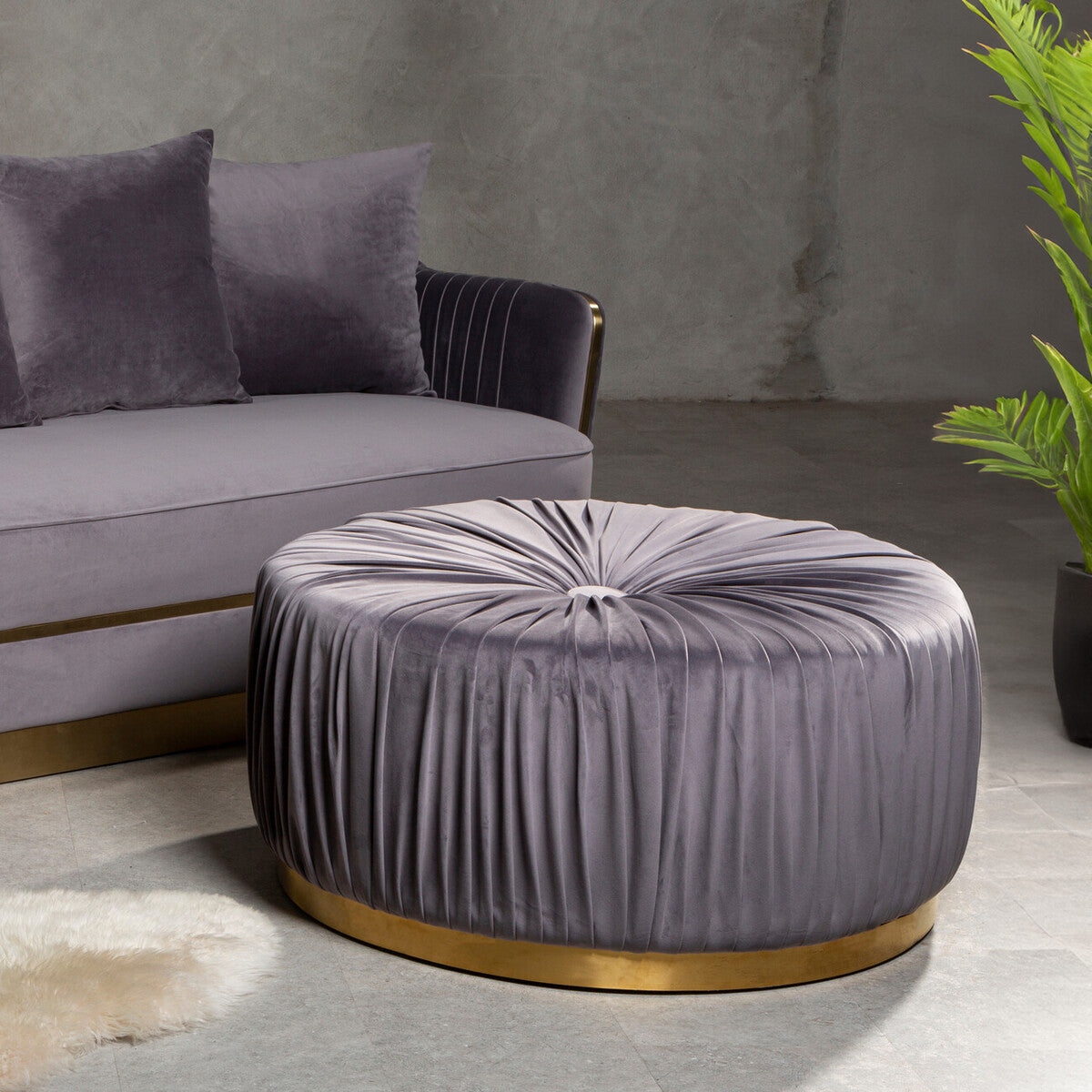 Round grey footstool with a tufted top and pleated fabric design, featuring a gold circular base, placed next to a grey sofa with matching cushions, set against a textured wall and greenery in the background