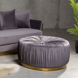 Round grey footstool with a tufted top and pleated fabric design, featuring a gold circular base, placed next to a grey sofa with matching cushions, set against a textured wall and greenery in the background