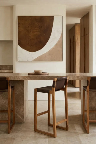 Brown leather bar chair with a teak wood frame, positioned at a modern kitchen counter. The background features a large abstract wall art piece in earthy tones, and the setting has a warm, neutral color palette with a minimalist aesthetic