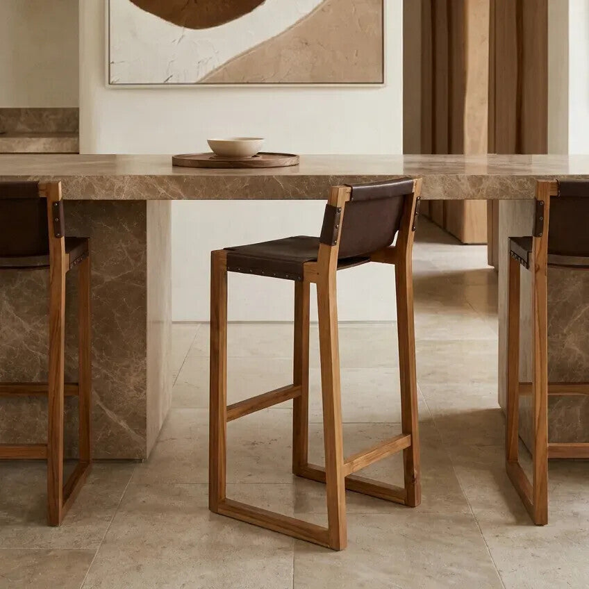 Brown leather bar chair with a teak wood frame, positioned next to a marble-topped bar table in a modern interior setting. The chair features a minimalist design with a sturdy, open frame and a comfortable seat. Soft lighting highlights the textures of the leather and wood, creating a warm and inviting atmosphere