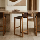 Brown leather bar chair with a teak wood frame, positioned next to a marble-topped bar table in a modern interior setting. The chair features a minimalist design with a sturdy, open frame and a comfortable seat. Soft lighting highlights the textures of the leather and wood, creating a warm and inviting atmosphere