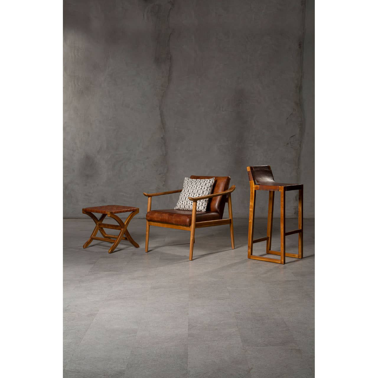 Brown leather bar chair with a teak wood frame, positioned next to a small wooden side table. The chair features a sleek design with a comfortable seat and backrest, while the side table has a minimalist structure. The background is a textured gray wall, enhancing the modern aesthetic of the furniture