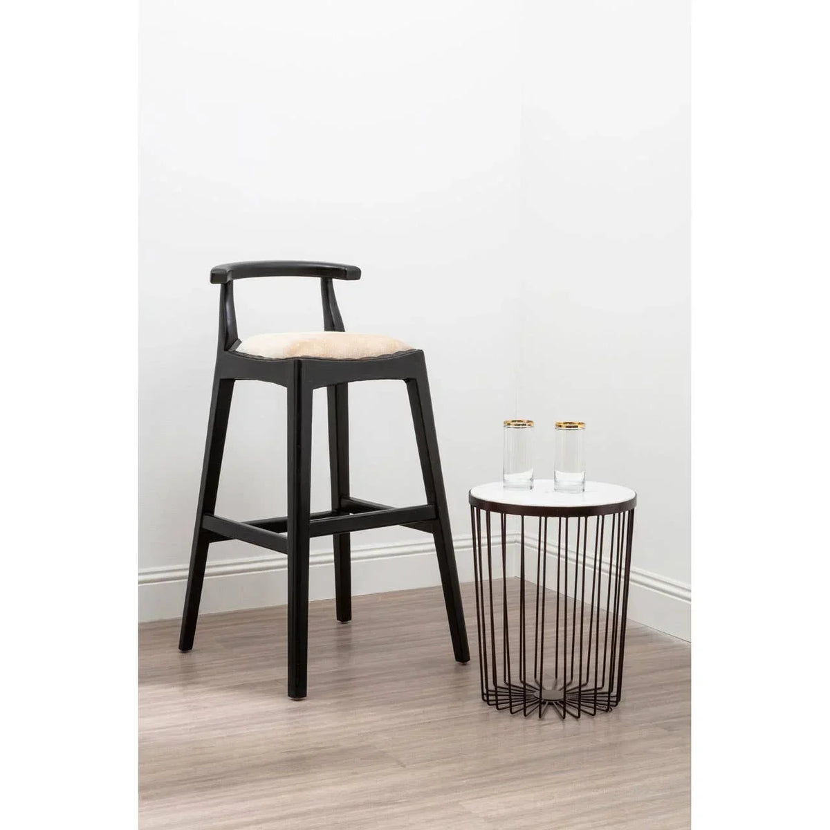 Black wooden bar stool with beige seat beside round wire side table and two glasses