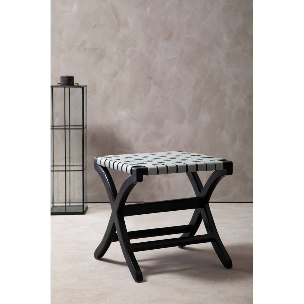 Woven grey leather stool with a black teak frame, featuring a cross-legged design and a textured seat, positioned against a neutral wall with a tall, minimalist lantern in the background