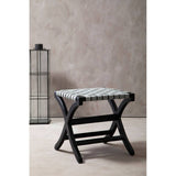 Woven grey leather stool with a black teak frame, featuring a cross-legged design and a textured seat, positioned against a neutral wall with a tall, minimalist lantern in the background