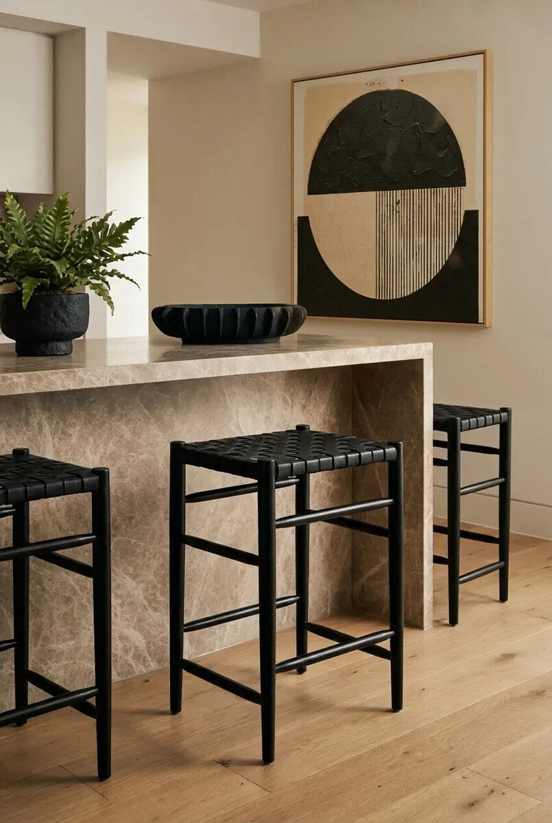 A set of three black woven leather barstools positioned around a light-colored stone countertop, with a decorative bowl and a potted plant on the counter. A large abstract wall art piece featuring geometric shapes is visible in the background, and the floor is made of light wood planks
