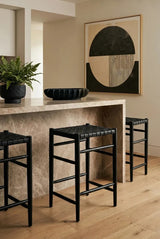 A set of three black woven leather barstools positioned around a light-colored stone countertop, with a decorative bowl and a potted plant on the counter. A large abstract wall art piece featuring geometric shapes is visible in the background, and the floor is made of light wood planks