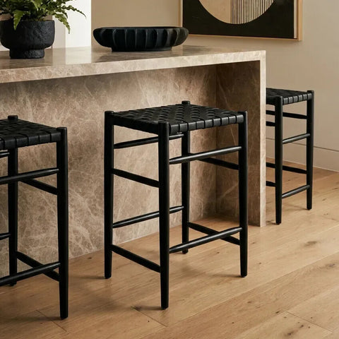 Three Kendari black woven leather barstools are positioned in front of a light-colored stone countertop, showcasing their sleek design and woven leather seats. The barstools feature a minimalist black frame with a simple, modern aesthetic, set against a backdrop of warm wooden flooring and a decorative bowl on the countertop