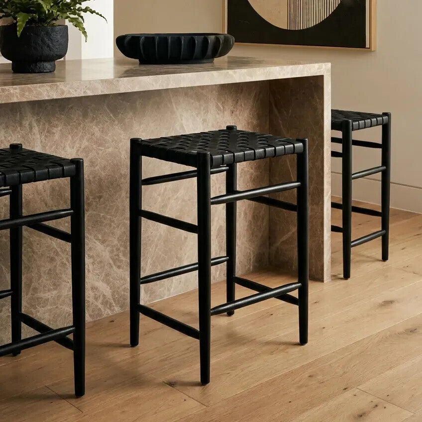 Three Kendari black woven leather barstools are positioned in front of a light-colored stone countertop, showcasing their sleek design and woven leather seats. The barstools feature a minimalist black frame with a simple, modern aesthetic, set against a backdrop of warm wooden flooring and a decorative bowl on the countertop