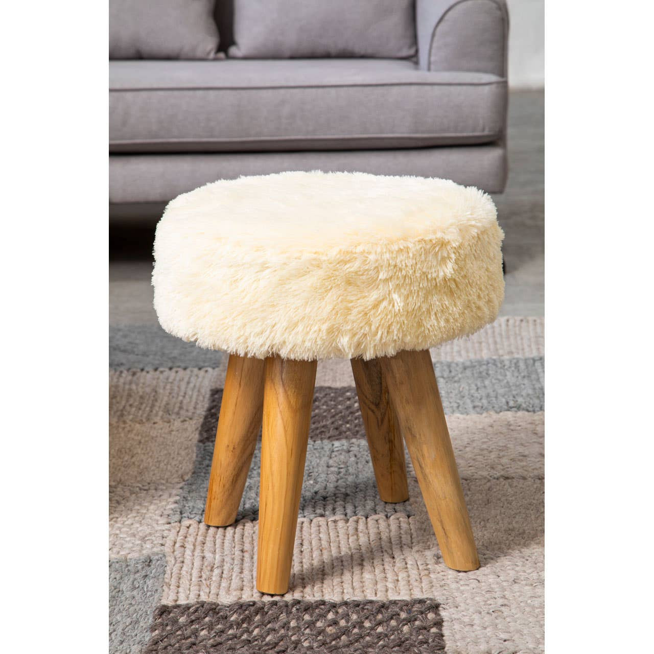 Cream faux fur stool with a round top and four tapered teak wood legs, placed on a patterned rug in a living room setting with a gray sofa in the background