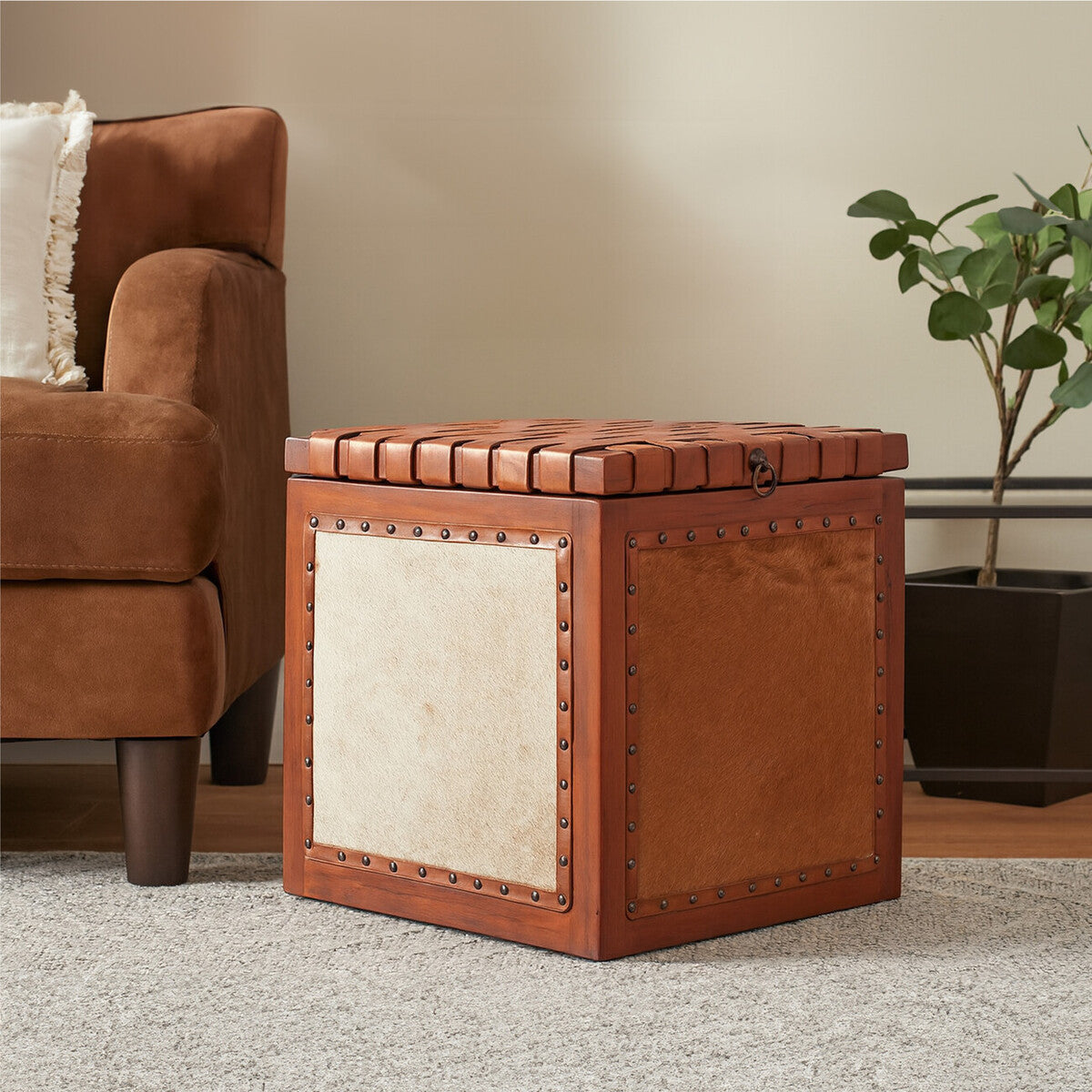 Square storage stool featuring a combination of hair-on leather and smooth leather, with a decorative studded trim along the top edge. The stool has a warm brown color and a contrasting light-colored panel on one side. It is placed on a light-colored rug next to a brown upholstered armchair and a small potted plant in the background