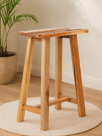 Kendari Teak Wood Stool with a natural finish, featuring a rectangular top and four sturdy legs, positioned on a round rug in a softly lit room with a potted plant in the background