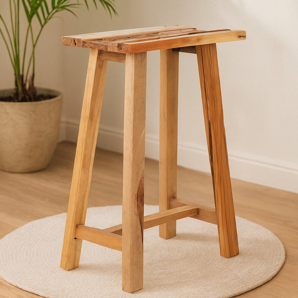 Kendari Teak Wood Stool featuring a natural wood finish, with a rectangular seat and four sturdy legs, placed on a round rug in a bright, minimalistic room with a potted plant in the background