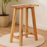Kendari Teak Wood Stool featuring a natural wood finish, with a rectangular seat and four sturdy legs, placed on a round rug in a bright, minimalistic room with a potted plant in the background