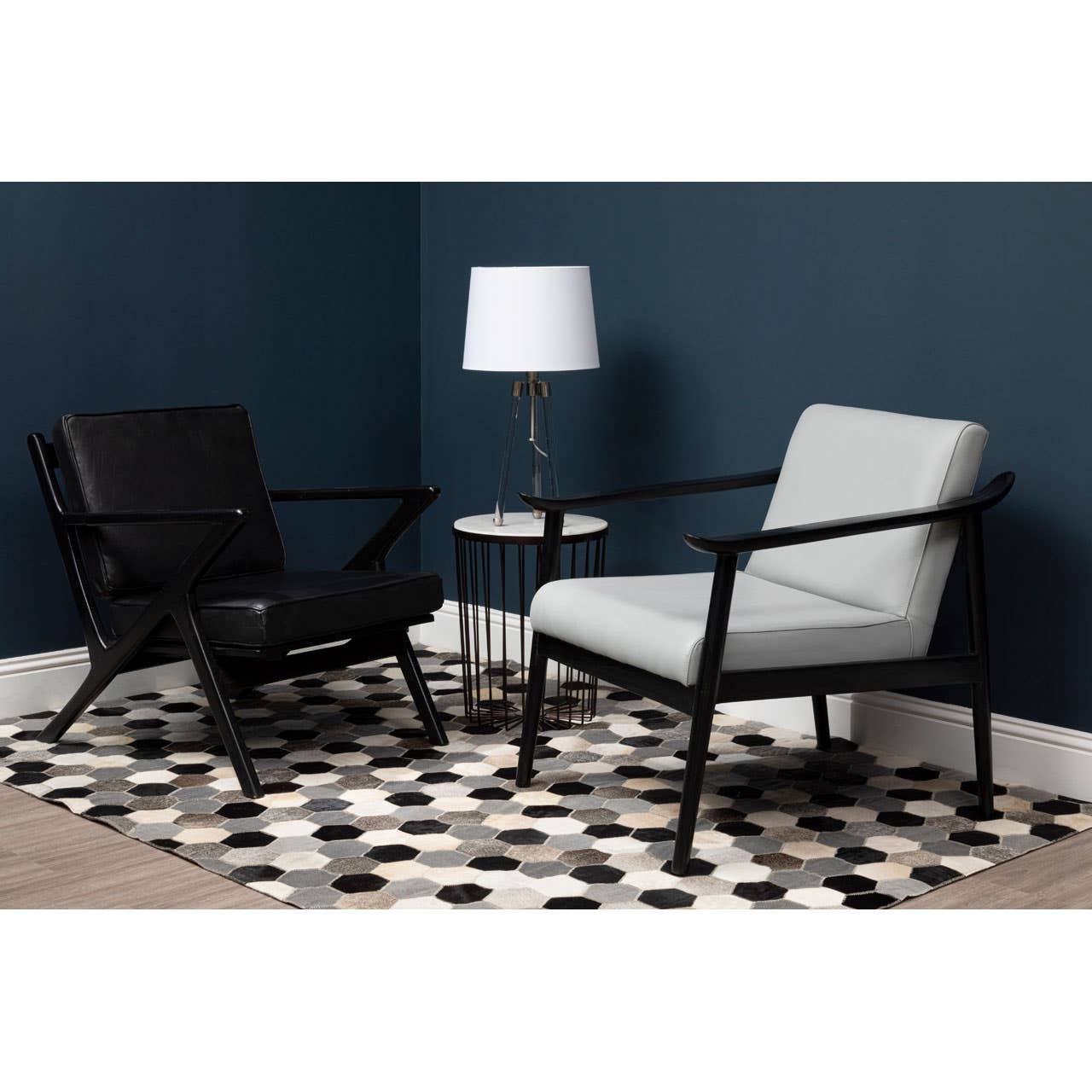 A black leather armchair with a teak wood frame is positioned next to a light gray armchair, both set against a dark blue wall. A small round side table with a white lamp is placed between the chairs, and the floor features a black and white patterned rug