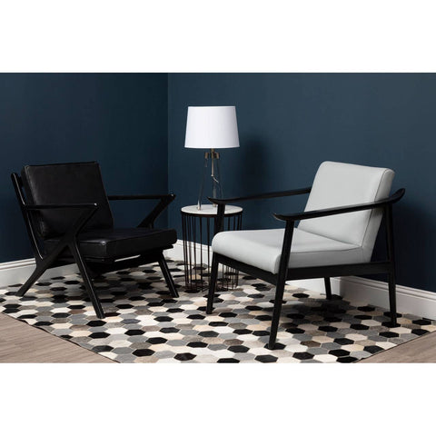 A black leather armchair with a teak wood frame is positioned next to a light gray armchair, both set against a dark blue wall. A small round side table with a white lamp is placed between the chairs, and the floor features a black and white patterned rug