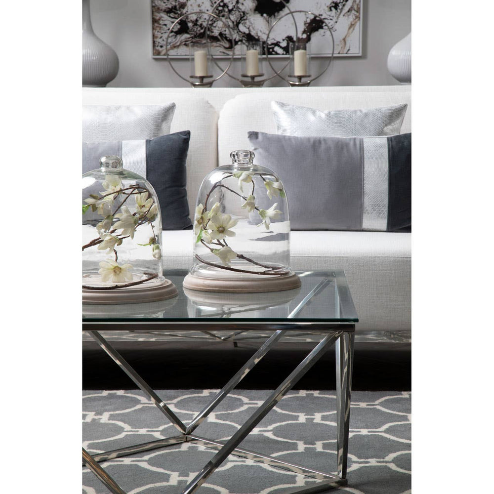 A stylish two-seater sofa in ivory and mink upholstery, positioned in a modern living room setting. The sofa features plush cushions and is complemented by a glass coffee table with decorative items, including two glass cloches and a patterned area rug beneath. The background showcases a piece of wall art and soft lighting, creating an inviting atmosphere